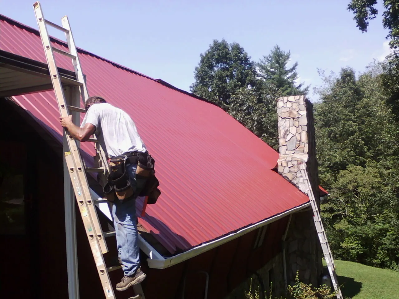 Roofer on a ladder beside a red metal roof and stone chimney — Soffit & Fascia Repair by Roofing Brothers in Quinebaug